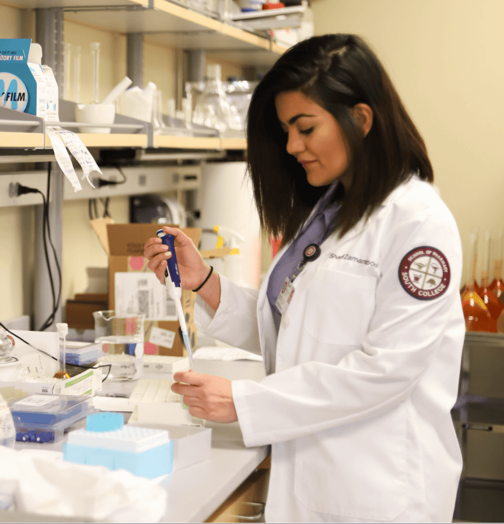 A Pharmacy student in a lab coat works in a laboratory, using a pipette to transfer liquid into a tray. Shelving with various lab supplies and equipment is visible in the background.