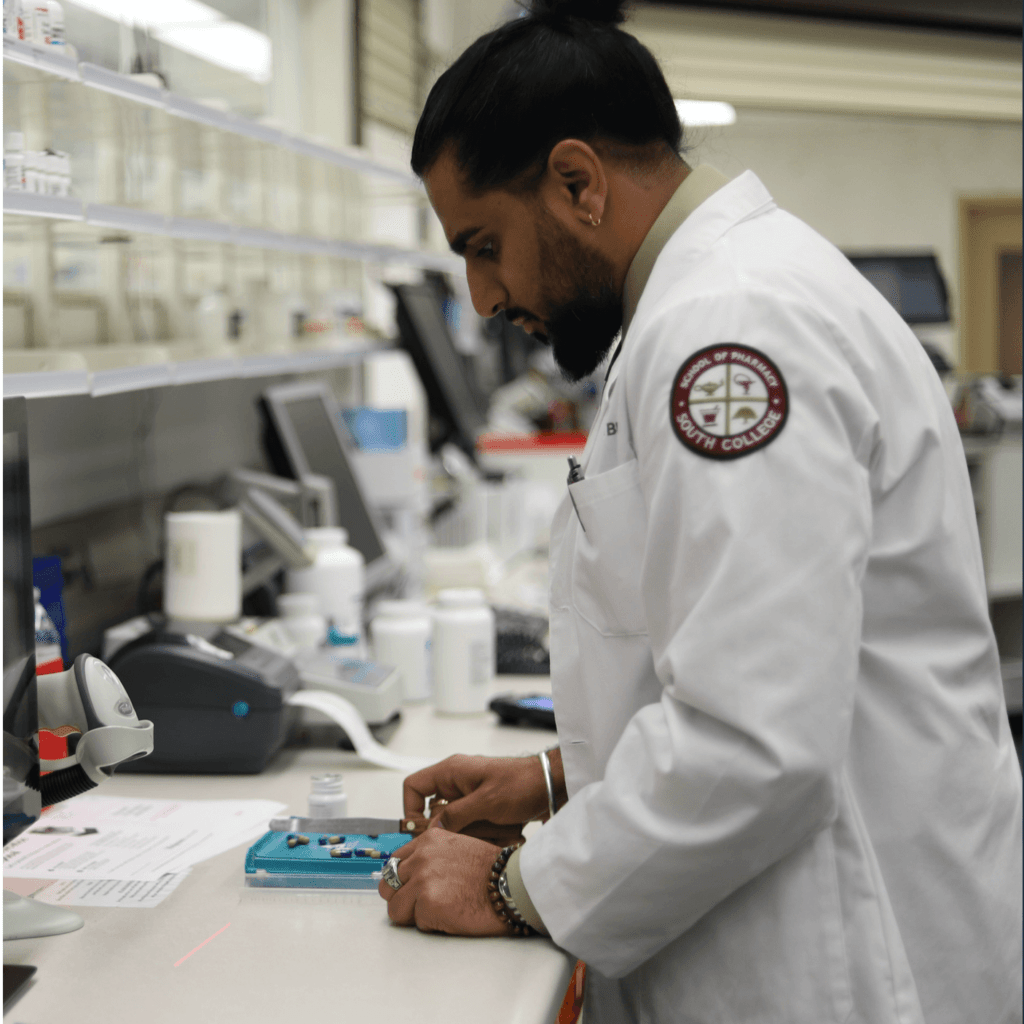 A Pharmacy student, wearing a white lab coat with a South College logo, is focused on sorting and counting pills on a tray at a pharmacy counter.