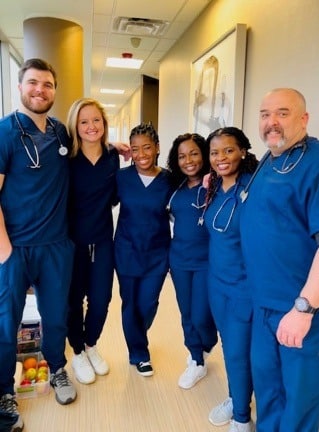 A group of six smiling nursing students, wearing navy blue scrubs and stethoscopes around their necks, stand together in a hallway. They include three men and three women, posing with their arms around each other.