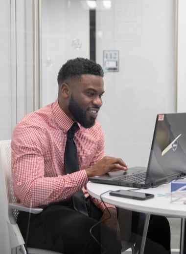 A man with a beard, wearing a red and white checkered shirt with a black tie, is sitting at a desk using a laptop. He appears to be engaged.