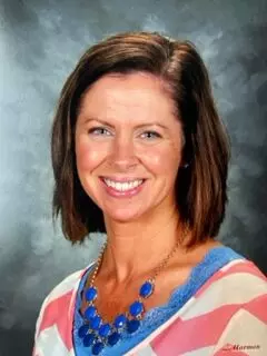 A woman with shoulder-length brown hair is smiling. She is wearing a top with pink and white stripes, a blue necklace, and blue lace detailing on the neckline. The background is a gray gradient.