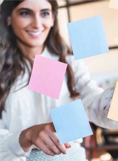 A person with long hair smiles and looks at colorful sticky notes suspended in the air in front of them. They appear to be organizing or arranging the notes. The background is slightly blurred, focusing on the person and the sticky notes.