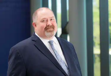 A man with a short beard, wearing a dark suit, a white shirt, and a patterned tie, stands in front of a modern building with large windows and vertical structures. He is smiling slightly and looking at the camera.