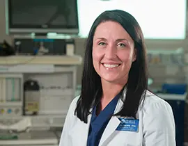 A woman with long dark hair, wearing a white medical coat and blue scrubs, smiles at the camera. Behind her is a medical office setting with various equipment and monitors.