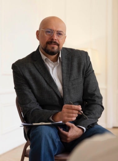 A man with glasses, dressed in a gray blazer and jeans, sits on a chair holding a pen and a notepad. He looks directly at the camera with a slight smile in a bright, softly lit room.