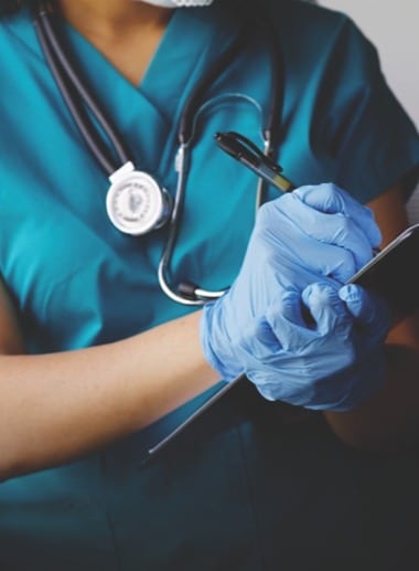 A nursing student wearing teal scrubs, a stethoscope around their neck, and blue gloves is holding a clipboard and pen, seemingly taking notes. The image focuses on their torso and hands.