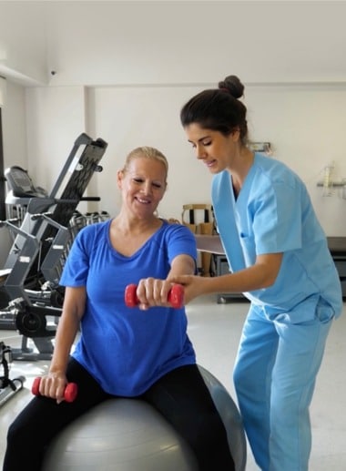 A physical therapy student in blue scrubs assists a smiling woman with arm exercises on a stability ball. The woman holds small red dumbbells in both hands. The scene takes place in a well-lit gym or rehabilitation center with exercise equipment in the background.