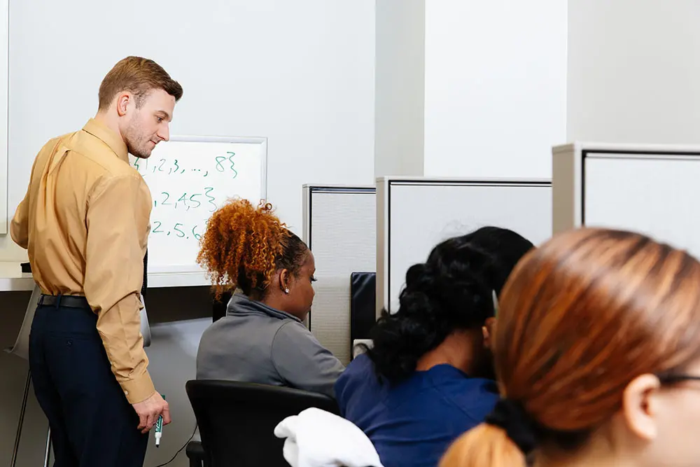 A man stands at a whiteboard with mathematical equations written on it, holding a marker. Several people are seated at desks in front of him, facing computer monitors, paying attention to his instruction.