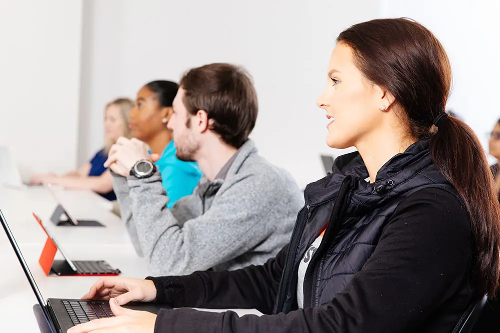 A group of students sit in a classroom setting, attentively looking towards the front. They have laptops open in front of them.