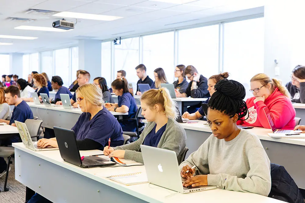 A diverse group of students is seated in a brightly lit classroom, working on laptops and notebooks.
