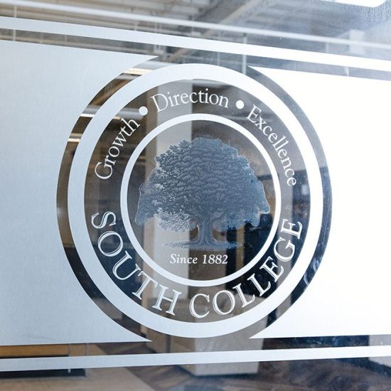 Glass window with a circular emblem for "South College" featuring a tree in the center. Text around the tree reads "Growth," "Direction," and "Excellence." Additional text says "Since 1882, recognized for accreditations and approvals." The emblem is etched in white, with some reflections visible on the glass.
