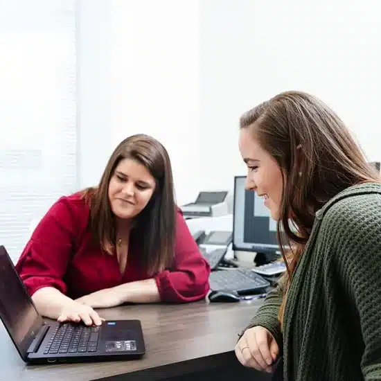 Two women sitting at a desk engage with a laptop. The woman on the left, in a red top, gestures towards the laptop screen, explaining something, while the woman on the right, in a green cardigan, listens attentively and smiles.