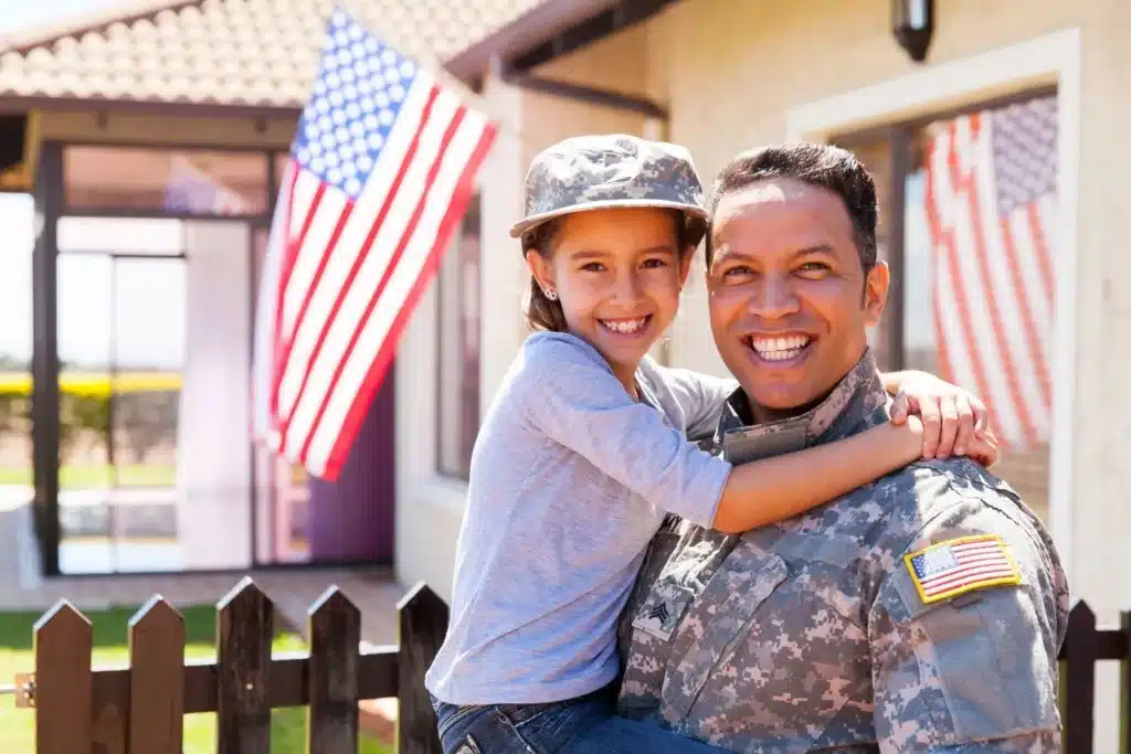 A smiling military person, dressed in a camo uniform with an American flag patch, is holding a child who is also wearing a camo cap.
