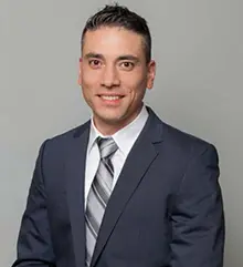 A man in a dark suit with a light gray striped tie and white shirt is smiling and facing the camera. He has short, dark hair and stands against a plain gray background.