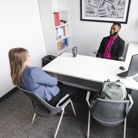 Two people are sitting across from each other in a modern office. One person, wearing a blue jacket, has their back to the camera.