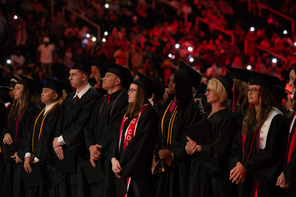 A group of graduates dressed in black caps and gowns stand in a row during their graduation ceremony. Some graduates hold diplomas and wear colorful cords and stoles.
