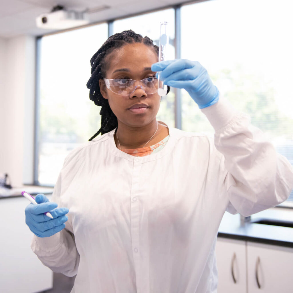 A student wearing goggles, gloves, and a white lab coat is focused on examining a test tube in a laboratory setting.