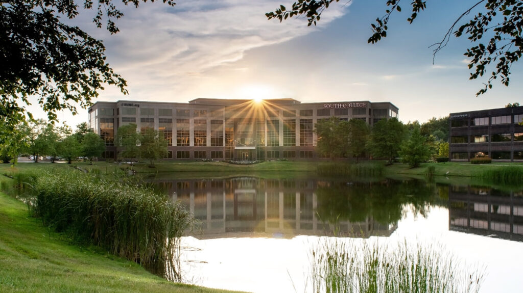 A modern office building with large glass windows sits in front of a pond, reflecting the structure and surrounding greenery. The sun is setting.