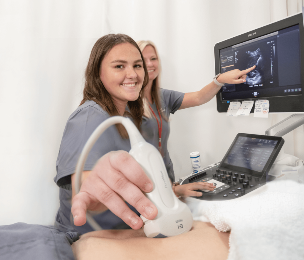 Two imaging science students in scrubs are performing an ultrasound. One holds the transducer and is smiling at the camera, while the other points at the ultrasound monitor.