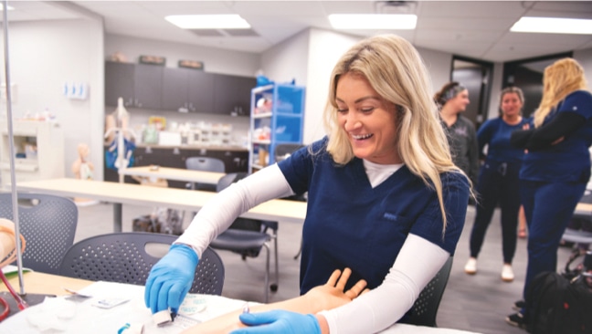 A woman in a blue scrub top smiles while practicing a medical procedure on a mannequin's arm in a classroom setting. She is wearing blue gloves.