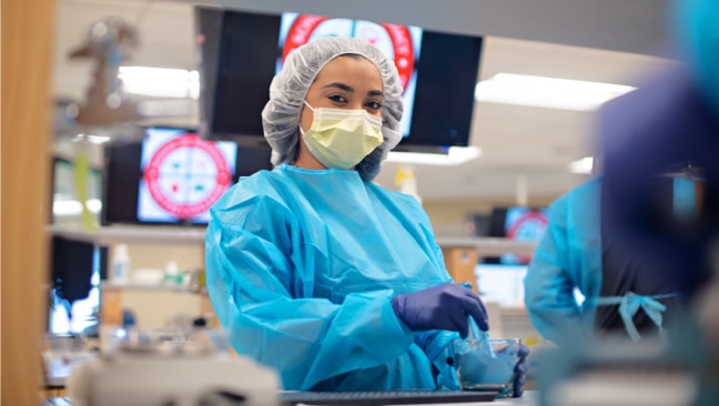A student wearing a blue surgical gown, hair net, face mask, and purple gloves stands in a medical facility.