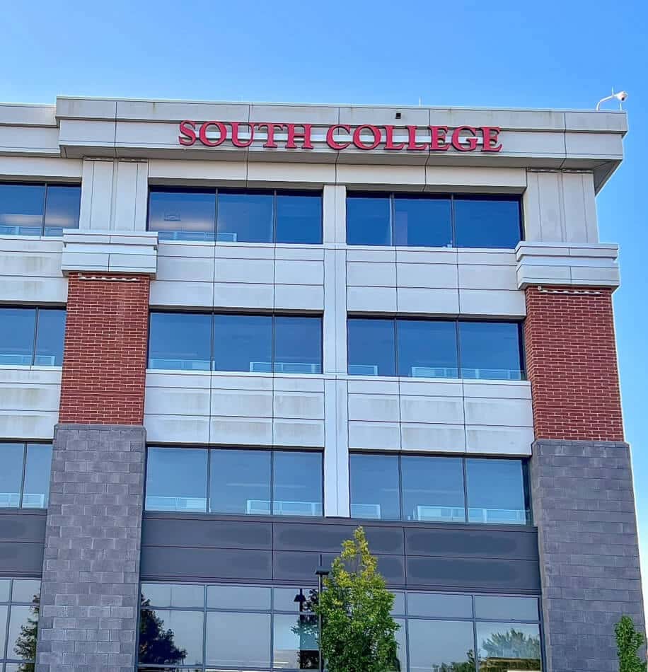 A multi-story building labeled "South College" at the top in red letters. The facade features a mix of brick, gray stone, and large glass windows.