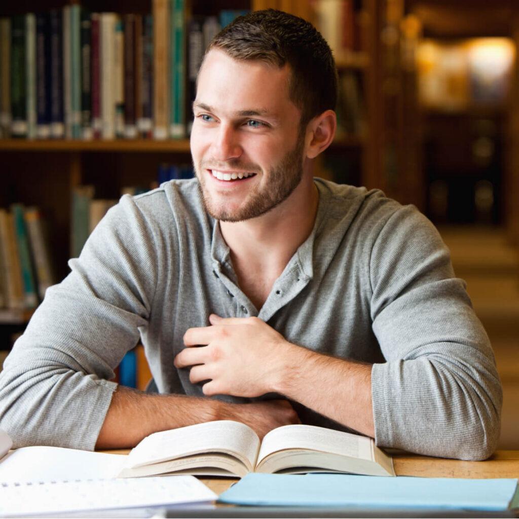 A smiling man with a beard and short hair sits at a wooden table in a library, wearing a gray long-sleeve shirt.