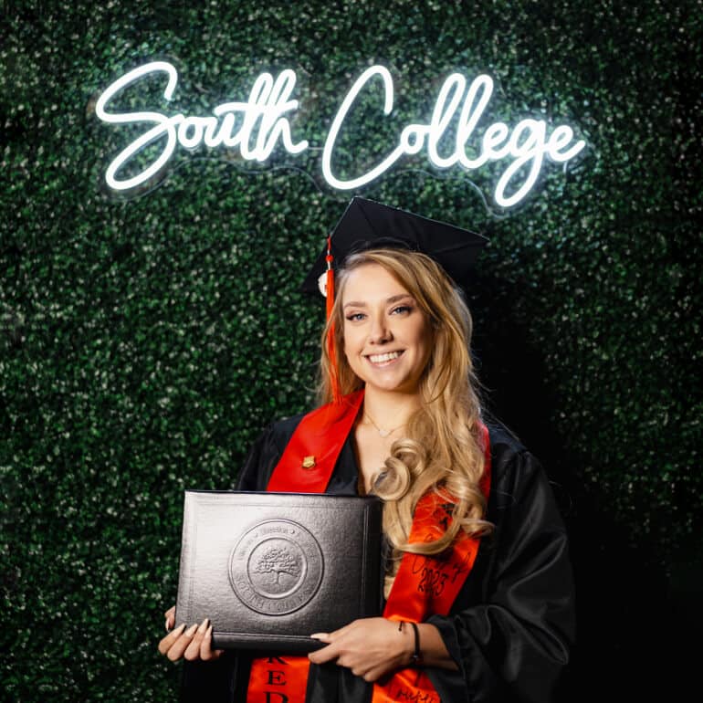 A young graduate in a black cap and gown with a red stole holds a diploma in front of a green leafy backdrop.