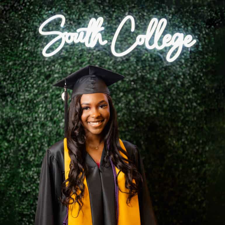 A graduate smiles in front of a backdrop with greenery and a neon sign that reads "South College."
