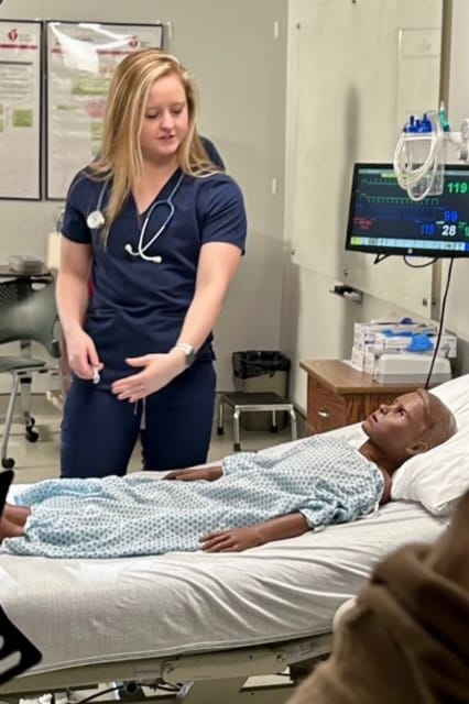 A Nursing student in navy scrubs with a stethoscope stands next to a hospital bed with a medical mannequin dressed in a hospital gown.