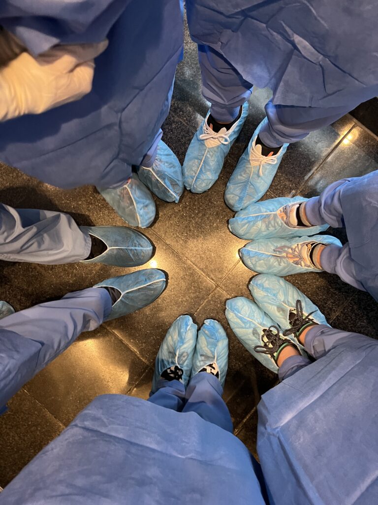 A group of people, seen from above, are standing in a circle wearing blue surgical scrubs and shoe covers.