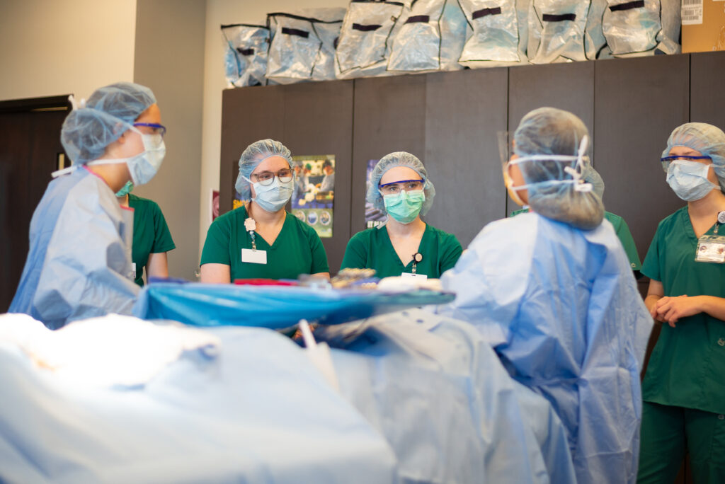 A group of medical professionals wearing green scrubs, surgical masks, hair nets, and protective eyewear stand around a patient draped in a blue surgical sheet, discussing the surgery. Medical supplies are visible in the background—a scene reminiscent of Tennessee Reconnect’s commitment to healthcare education.