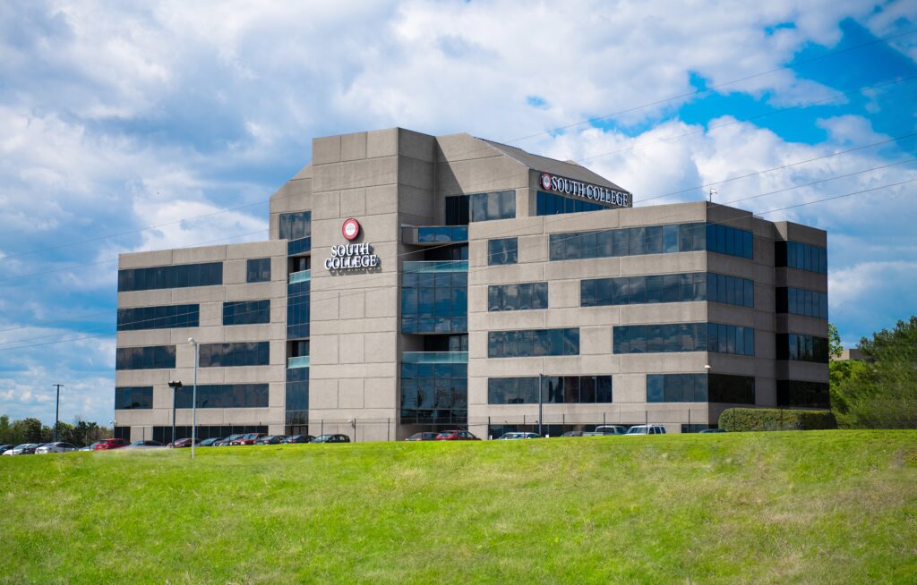 A modern, multi-story building with a facade of reflective glass and concrete. The building displays the name "South College" on two signs. Several cars are parked in the lot in front, and a green grassy area is visible in the foreground. The sky above is partly cloudy.