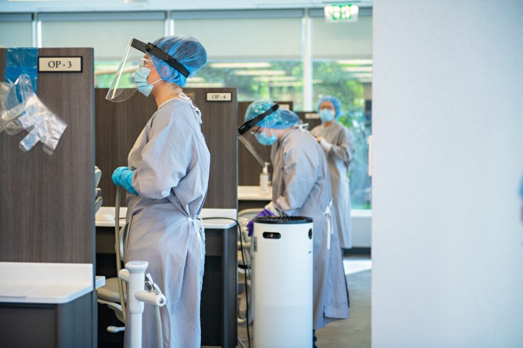 Dental students in protective gear, including face shields, masks, gowns, and gloves, attend to patients in a clinic. They are in separate cubicles labeled OP-3 and OP-4, engaging with equipment. One student is visible in the background.