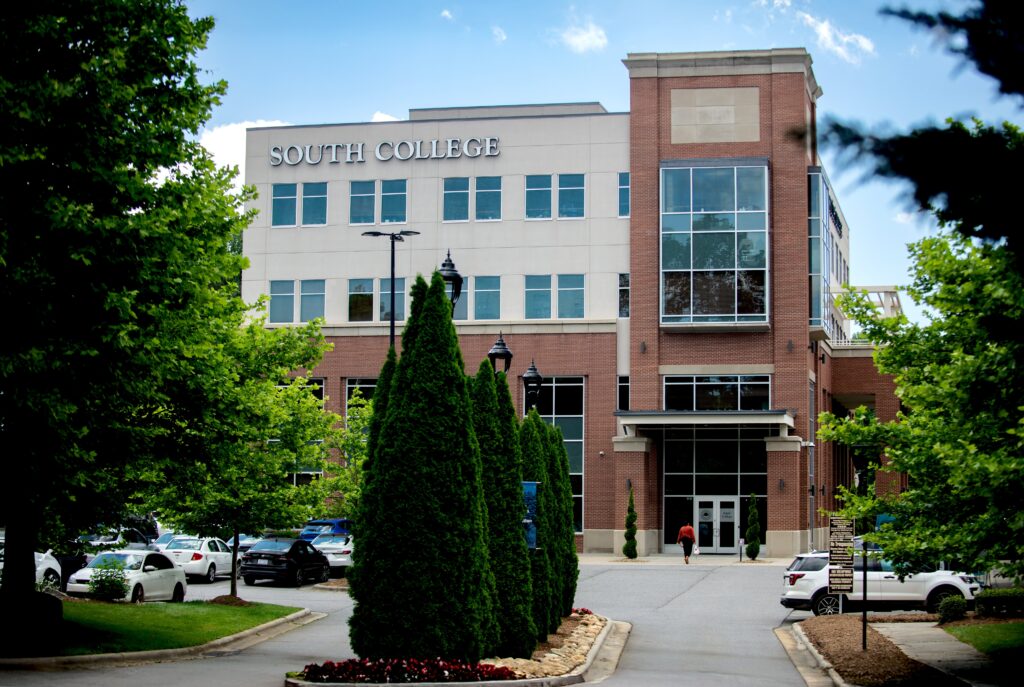A modern brick and glass building with “South College” signage, surrounded by trees and parked cars, on a bright day. A sidewalk lined with tall shrubs leads to the main entrance.